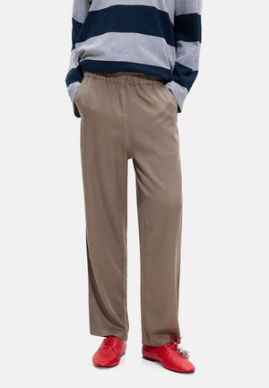 Osoba ubrana w luźne brązowe spodnie, szaro-granatową bluzkę w paski z długim rękawem oraz jasnoczerwone buty z ozdobnymi zawieszkami na jednej sznurówce.