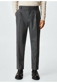 Homme portant un pantalon gris sur mesure, un pull côtelé crème et des bottes en cuir noir, debout les mains détendues le long du corps.