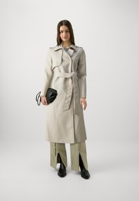 Beige trenchcoat met een ceintuur in de taille, voorzien van dubbele knopen en schouderepauletten, gecombineerd met groene broek en zwarte laarzen.