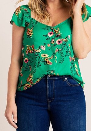 Kobieta w zielonej bluzce z krótkim rękawem w kwiatowy wzór i guziki, w połączeniu z ciemnoniebieskimi, wysokim stanem jeansami.