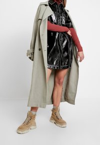 Ljusgrön trenchcoat över en svart glansig klänning. Beige chunkyskor med snören. Röd långärmad topp under. Minimala detaljer.