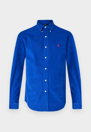 Chemise en velours côtelé bleu à manches longues, col boutonné, boutons blancs et petit logo rouge sur la poitrine. Texture lisse, coupe ajustée.