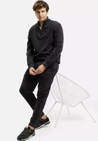 Młody mężczyzna w czarnej bluzie z krótkim zamkiem i dresowych spodniach siedzi na białym krześle o stalowej konstrukcji, w czarnych sneakersach, na białym tle.