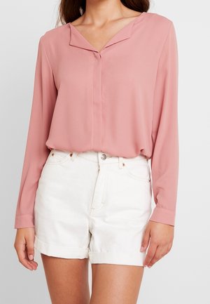 Kvinde iført en lyserød bluse med lange ærmer, der er stoppet ned i hvide shorts med høj talje og opbukket kant, stående foran en ensfarvet baggrund.