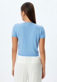 Maglione azzurro chiaro a maniche corte con orlo e colletto a coste. Texture morbida, design semplice. Indossato con pantaloni bianchi su misura, mostrando la vista posteriore.