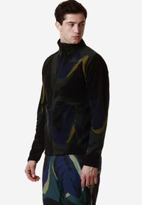 Giacca in pile nera con motivi astratti verdi, blu e gialli; presenta una chiusura con zip e tasche laterali.