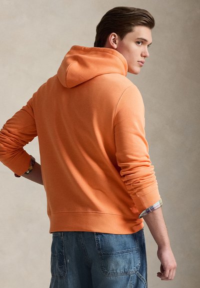 Πορτοκαλί φούτερ με απαλό ύφασμα, που διαθέτει κουκούλα, ριμπ μανσέτες και τσέπη καγκουρό. Φοριέται με τζιν σορτς και μια καρό πουκάμισο από κάτω.