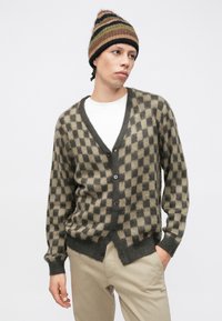 Cardigan a quadri marrone e beige in morbido maglione, caratterizzato da una scollatura a V profonda, chiusura con bottoni e polsini a costine, abbinato a un cappello a maglia a righe.