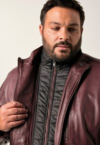 Burgundská kožená bunda s černou prošívanou vnitřní vrstvou, vybavená vysokým límcem a zipy. Kůže vypadá hladce, zatímco vnitřní textura je vycpaná.