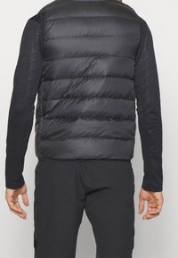 Svart puffväst med horisontella quiltade sektioner, kombinerad med en långärmad svart topp. Materialet verkar smidigt och lätt.