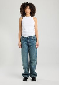 Witte mouwloze tanktop gecombineerd met losse, mid-blauwe spijkerbroek en zwarte chunky schoenen, met een rechte pijp en vervaagde textuur.