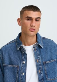 Denim jakna tamnoplave boje s klasičnom kragnom, prednjim džepom, metalnim dugmadi i šivenim detaljima. Nosi se preko bijele majice.