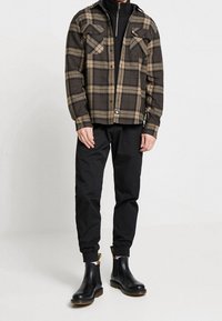 Geruite jas in bruin en beige, met twee borstzakken, gecombineerd met zwarte tapse broeken en zwarte chelsea boots met gouden accenten.