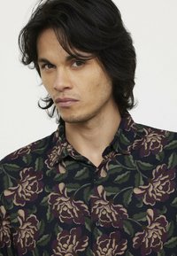 Korte mouwen overhemd met knopen in donkere stof met een bloemenpatroon in tinten beige, groen en bordeauxrood, met een camp collar en zichtbare knoppen.
