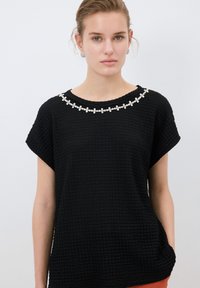 Top nero a trama con maniche corte, caratterizzato da un motivo decorativo di perline floreali bianche intorno allo scollo. Design moderno e vestibilità morbida.