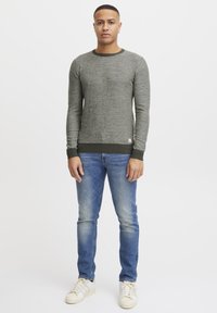 Gray knitted sweater with a crew neckline and ribbed cuffs, paired with blue jeans and white sneakers. Model stands against a plain background.