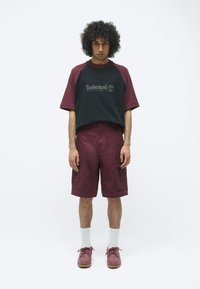 Bordeauxrode shorts en een zwart raglan t-shirt met een Timberland-logo. Het shirt heeft korte mouwen; de outfit bevat bruine schoenen en witte sokken.