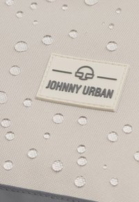 Beige táska texturált felülettel, vízcseppekkel és egy téglalap alakú, krémszínű címkével, amelyen sötét betűkkel az áll: "JOHNNY URBAN".