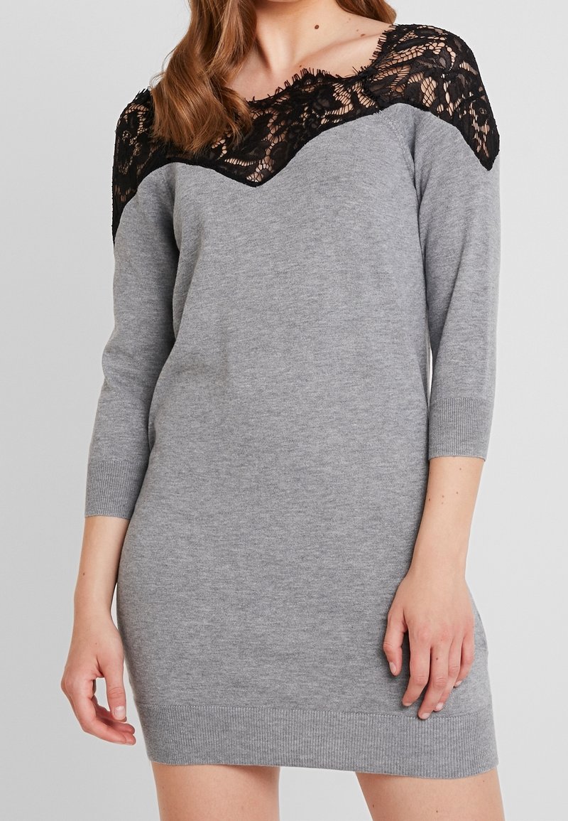 Femme portant une robe en maille grise avec des manches trois-quarts et un détail en dentelle noire sur les épaules et le décolleté.