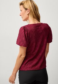 Mørkerød bluse med puffede ærmer, der har et blomster-krone design og åben syning. Blød tekstur, rund halsudskæring og dekorative kantdetaljer.