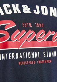 Camisa de algodón navy con texto gráfico en blanco y rojo, que incluye "SUPER" y "NORMAS INTERNACIONALES", con detalle del año de establecimiento.