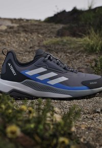 Γκρι και μπλε παπούτσι τρεξίματος Adidas Terrex trail σε πετρώδες έδαφος με λευκές ρίγες και κείμενο RAIN.RDY στην προστασία δακτύλων.