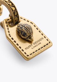 Accessorio in ottone con un gancio a forma di uccello, superficie texturizzata e incisa "Kurt Geiger London" su un'etichetta rettangolare. Accentuato da pietre.