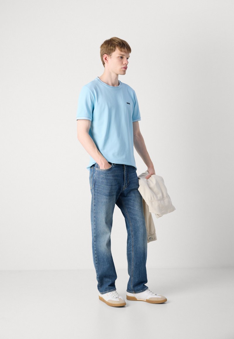 T-shirt bleu clair à manches courtes avec des finitions contrastées, jean foncé, baskets blanches, tenant une veste claire. Look simple et décontracté.