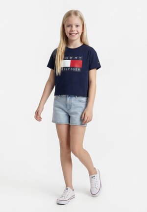 Ξανθό κορίτσι στέκεται και χαμογελά φορώντας ένα σκούρο μπλε T-shirt Tommy Hilfiger, ανοιχτόχρωμα τζιν σορτς και λευκά αθλητικά παπούτσια σε λευκό φόντο.