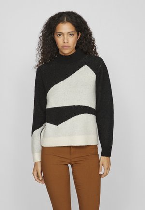 Suéter de punto con cuello alto, patrón geométrico en blanco y negro y dobladillo acanalado. Combinado con pantalones ajustados marrones.