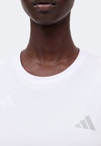 Gros plan sur le cou et la partie inférieure du visage d'une personne portant un t-shirt blanc Adidas avec un logo gris sur la poitrine, sur un fond uni.