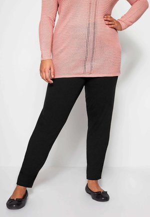 Vrouw die een roze gebreide top met lange mouwen draagt en zwarte rechte broek met zwarte ballerina's, staand met de hand op de heup.