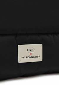 Sac rembourré noir avec une surface texturée, doté d'une étiquette crème. L'étiquette affiche "UNIO HAMBURG X LES VISIONNAIRES" en texte noir et rouge.