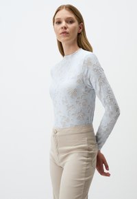 Top a maniche lunghe in pizzo azzurro chiaro con motivo floreale, abbinato a pantaloni beige a vita alta con un unico bottone frontale e tasche laterali.