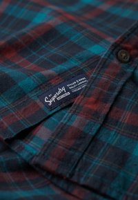 Rood, groen en blauw geruit overhemd met een zachte textuur. Voorzien van een stoffen label met het "Superdry" logo en bruine knoopaccenten.