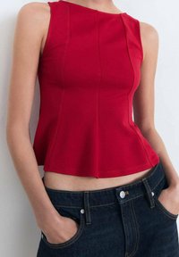 Red sleeveless top with a fitted bodice and peplum hem; smooth fabric with seam detailing. Paired with dark denim jeans.
