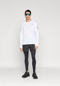 Λευκό αθλητικό πουκάμισο με μακριά μανίκια και γκρι ρίγες, μαύρο σορτς, μαύρο κολάν και μαύρα αθλητικά παπούτσια. Το μοντέλο φορά γυαλιά ηλίου με γαλάζιο φακό.