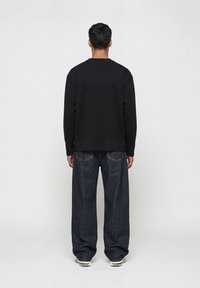 Άνδρας που στέκεται με την πλάτη γυρισμένη, φορώντας μαύρο μακρυμάνικο πουκάμισο, φαρδιά σκουρόχρωμα μπλε τζιν και λευκά αθλητικά παπούτσια σε απλό φόντο.