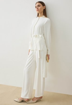 Kobieta stojąca przy białej ścianie, ubrana w biały, długorękawowy top z paskiem, szerokie białe spodnie, beżowe buty na obcasie typu slingback, trzyma małą kopertówkę.