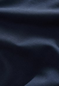 Laivastonsininen kangas, jossa on sileä pinta, hienovarainen kiilto ja valoa heijastavat kuviot, näyttää rypistyneeltä ja laskeutuneelta.