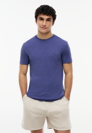 Jeune homme aux cheveux bouclés portant un T-shirt bleu à manches courtes et un short beige, les mains dans les poches, debout devant un fond blanc.