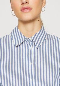 Camicia a righe blu e bianche con colletto, frontale con bottoni e tessuto dalla texture liscia. Presenta un design classico con righe orientate verticalmente.