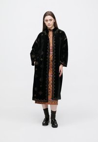 Ilgas juodas aksominis paltas, puoštas sudėtingais aukso siuvinėjimais ir dekoratyviniais mygtukais, vilkimas ant raštuotos suknelės ir derinamas su tamsiais batais.