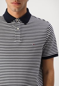 Bărbat care poartă un tricou polo cu dungi orizontale bleumarin și albe, cu guler bleumarin și un logo mic pe piept.