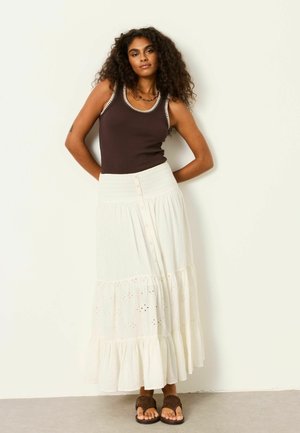 Frau mit lockigem Haar, die ein dunkelbraunes ärmelloses Oberteil, einen weißen, gestuften Maxirock und braune Sandalen trägt und vor einer schlichten Wand steht.