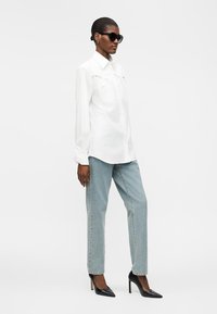 Femme debout portant une chemise blanche oversize à boutons, un jean bleu clair, des talons hauts noirs et de grandes lunettes de soleil noires sur un fond uni.