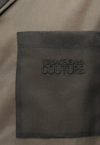 Μαύρη υφασμάτινη τσέπη με κεντημένο τοποθετημένο μαύρο νήμα κείμενο "VERSACE JEANS COUTURE" σε καφέ ένδυμα.