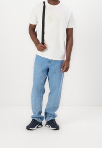 Man med vit grafisk T-shirt, ljusblå jeans med sydda fickor, svarta sneakers och bär en svart rem över axeln.