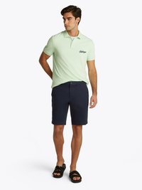 Polo de cor verde claro com calções escuros de marinha. A polo tem colarinho e um logótipo bordado no peito. Usado com sandálias pretas.