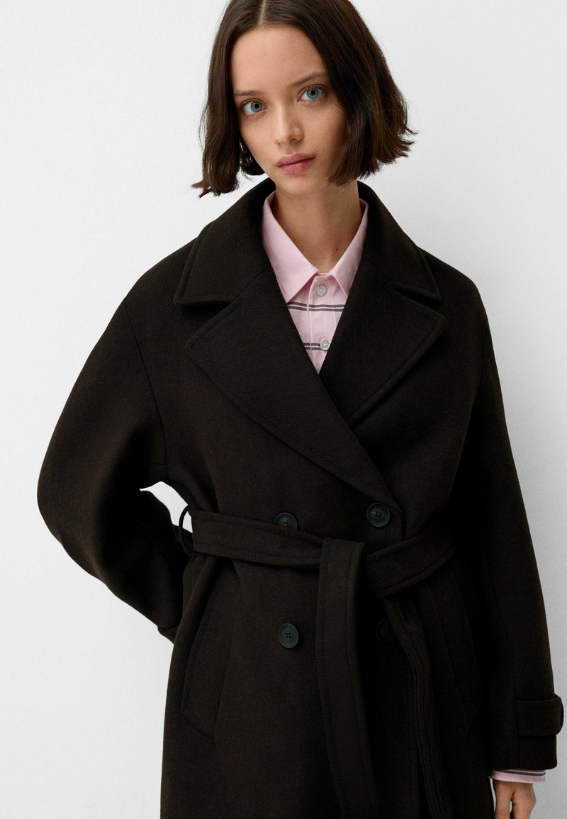 Ciemnobrązowy wełniany płaszcz o podwójnym zapięciu, z dużymi klapami i dopasowanym paskiem; posiada akcenty guzikowe oraz boczne kieszenie.